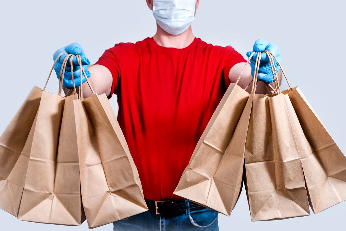 Safe Delivery. A Courier In A Red Uniform And Protective Mask And Gloves Holds A Large Order, Many Paper Bags, Contactless Delivery Food In Quarantine. Donation Of Volunteers. Zero Waste.