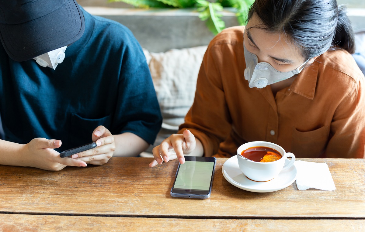 Two Womans In Protective Mask Prevent Covid 19 Using Cell Phone And Drinking Lemon Tea In Cafe.